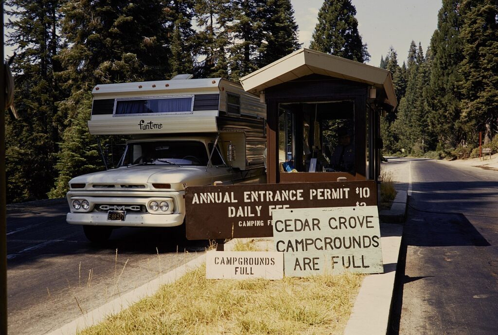 A vintage GMC camper parked at a park entrance, with signs indicating camping fees and that Cedar Grove campgrounds are full.
