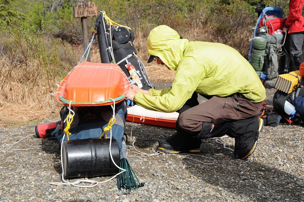 A person wearing a bright yellow jacket secures gear with ropes near an orange canoe at a trailhead marked "McKinley Bar Trail."