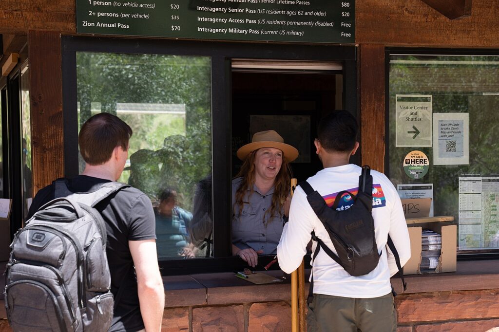Two visitors with backpacks approach a park entrance kiosk displaying pass prices and information about services.