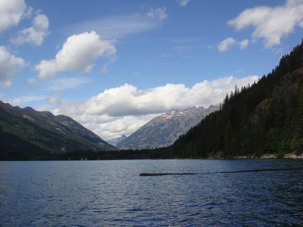 Serene mountain lake scene with lush green forests, majestic peaks, and a blue sky dotted with fluffy clouds.