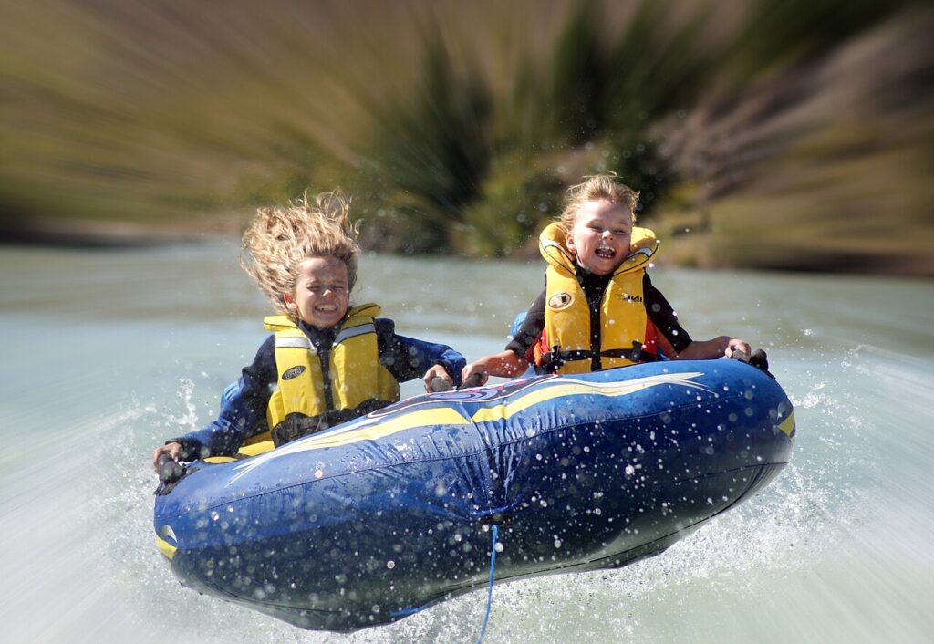 Two children in life jackets soar above the water on a blue inflatable tube, splashing and enjoying a thrilling ride.