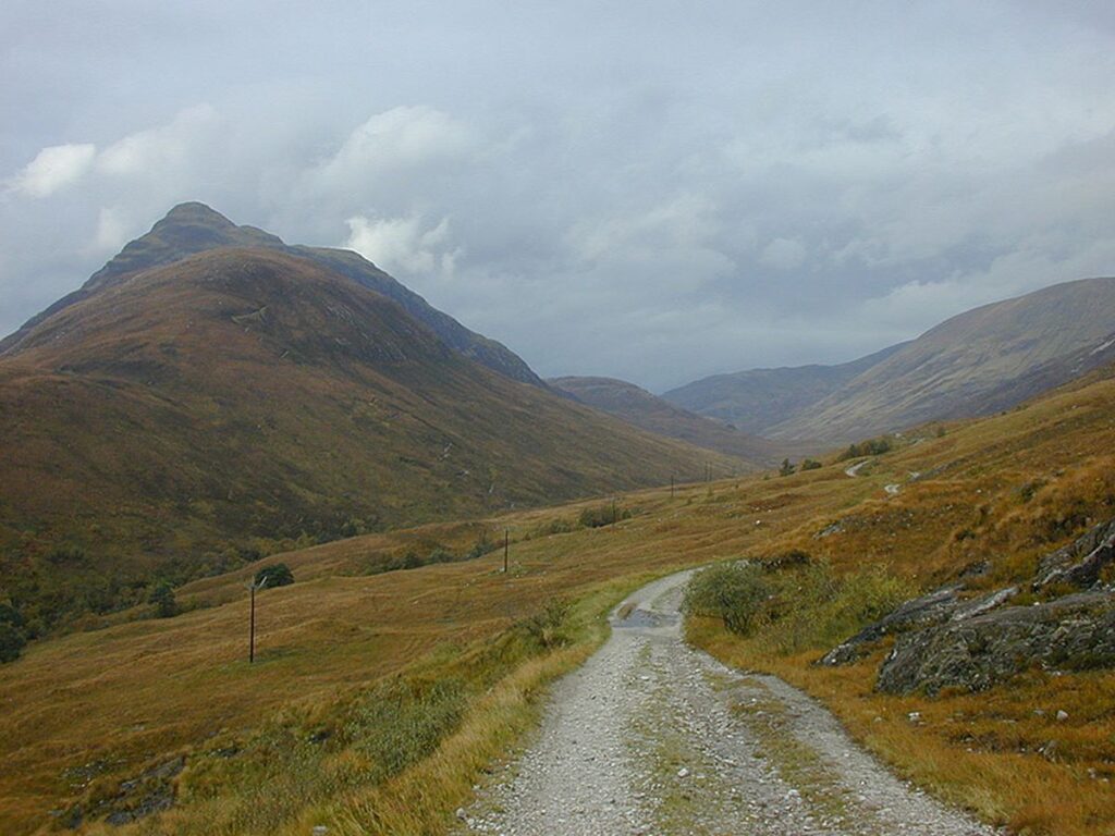 A winding gravel road cuts through a rugged valley surrounded by rolling hills under a cloudy sky in a serene mountain landscape.
