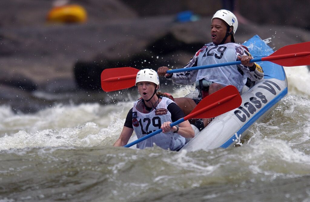 Two individuals paddle a blue raft through white water rapids, surrounded by splashing waves and rocky terrain.