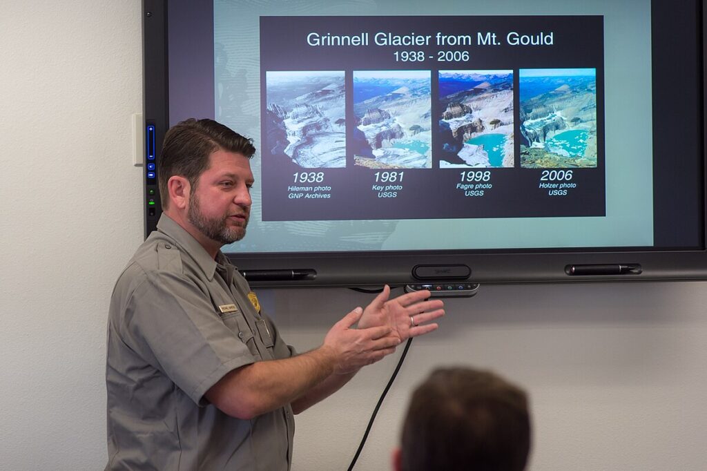 A presenter discusses Grinnell Glacier's changes over time, showcasing images from 1938, 1981, 1998, and 2006 on a digital screen.