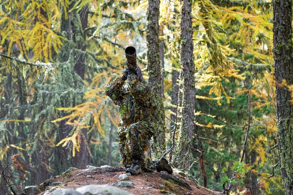A camouflaged photographer stands on a rock in a forest, aiming a camera through a large lens amidst vibrant autumn foliage.