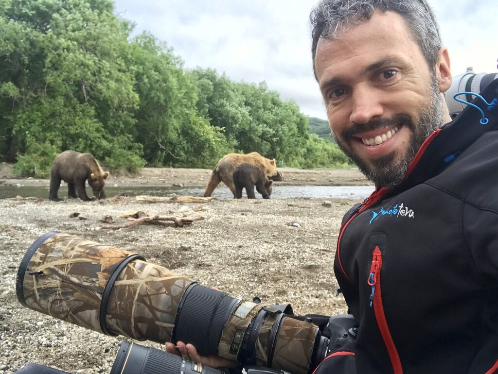 A photographer sits on a gravel bank, capturing images of three brown bears foraging by a river amid lush greenery.