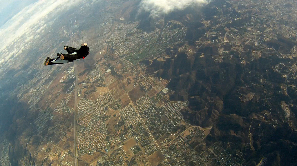 A skydiver soaring through the air above a sprawling landscape of houses and hills, clouds drifting above them.