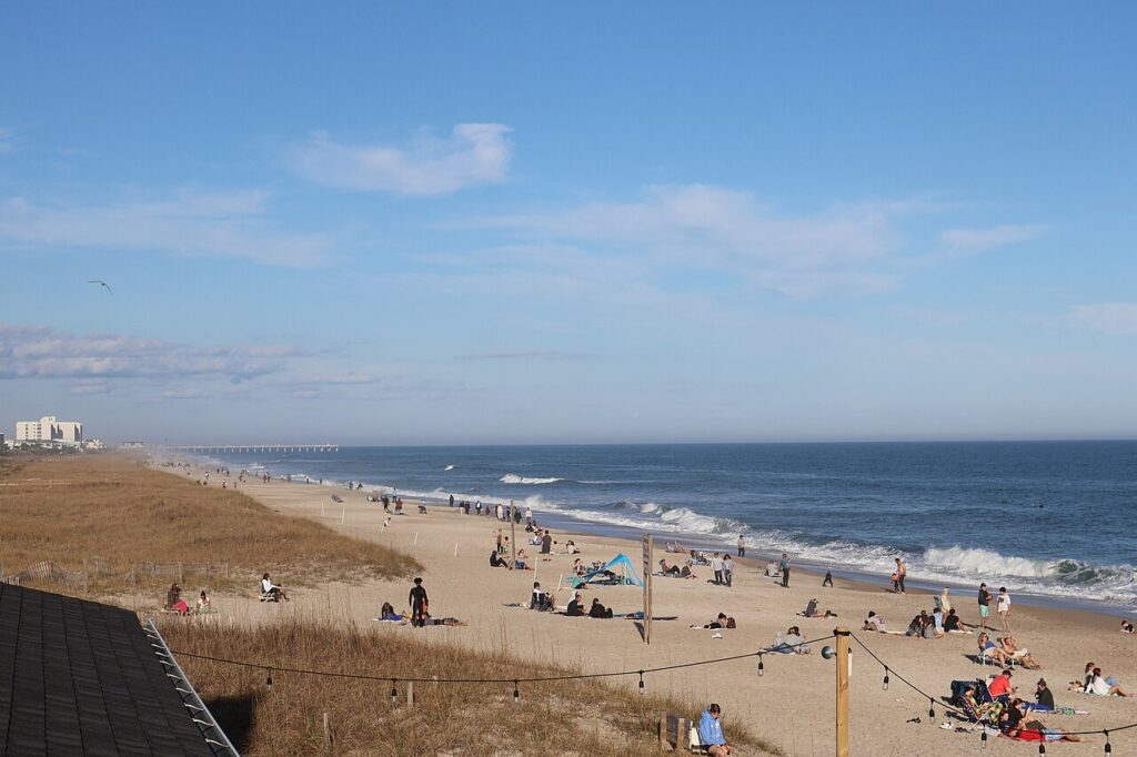 A sunny beach scene with people lounging, walking, and playing along the shore, with waves lapping at the sand under a clear blue sky.
