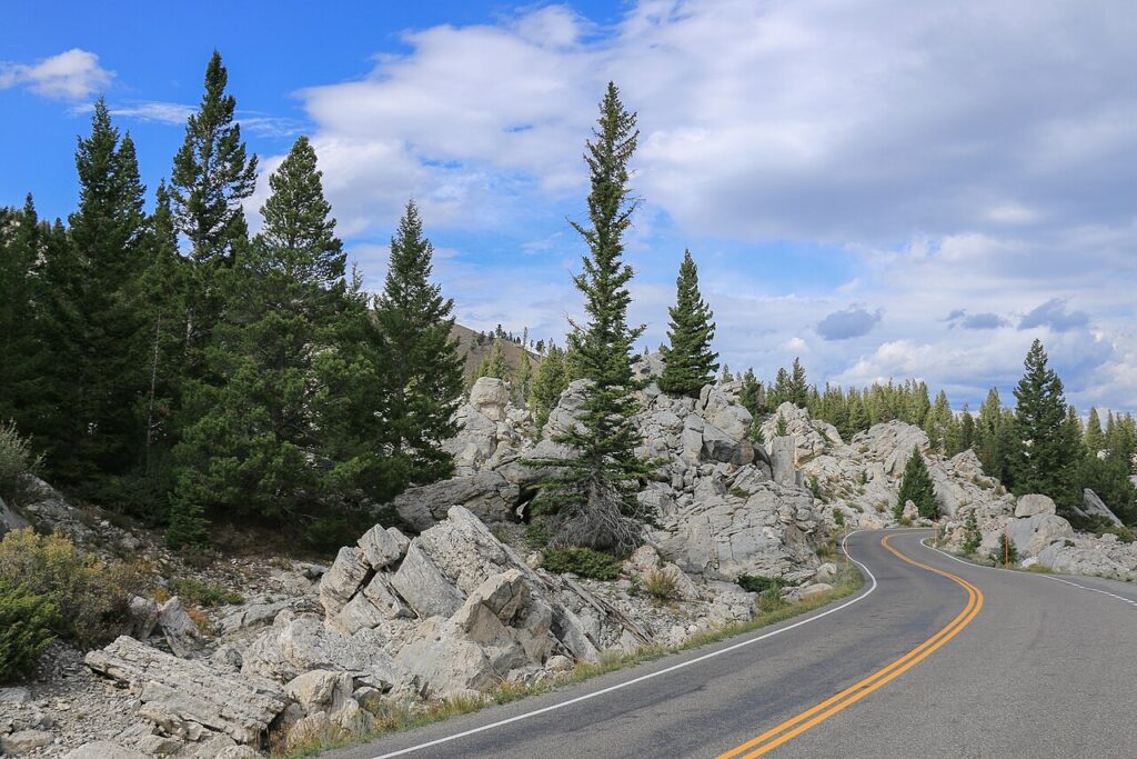 A winding road curves through rocky terrain and tall evergreen trees under a partly cloudy sky.