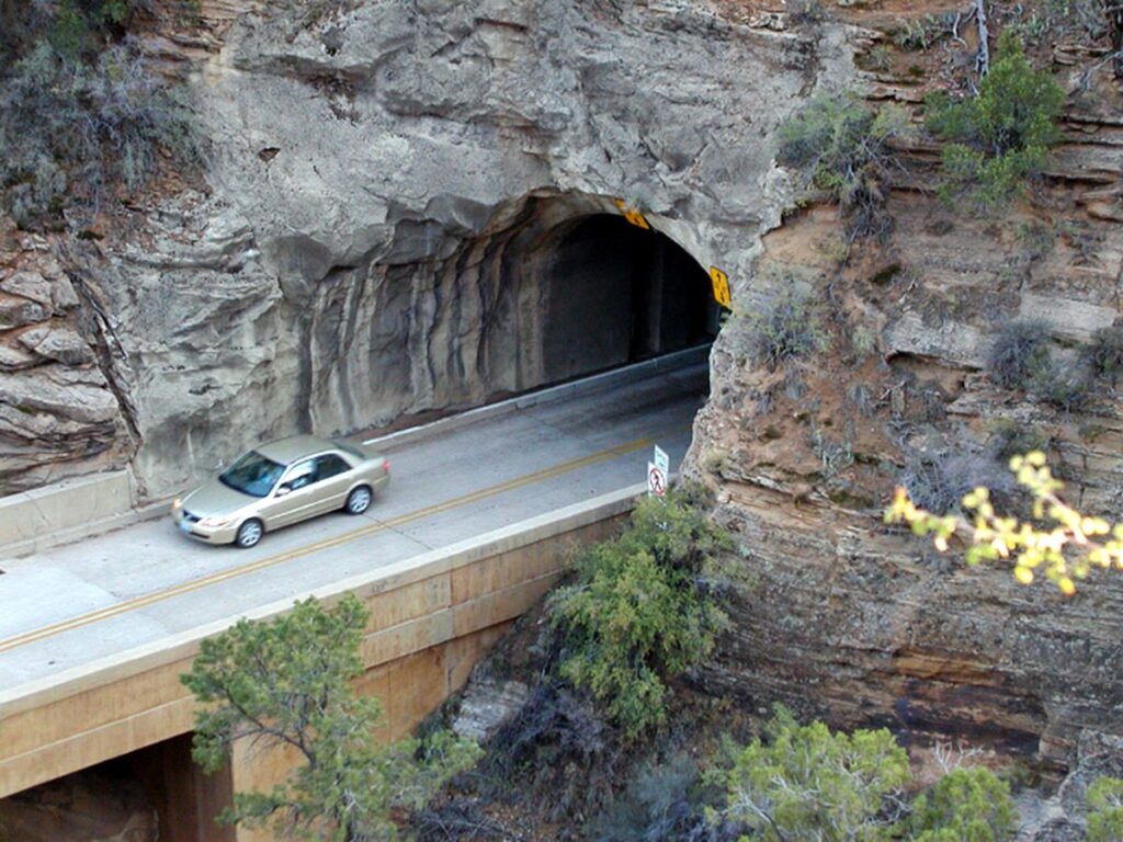 A silver car drives through a rocky tunnel on a two-lane road, surrounded by rugged terrain and sparse vegetation.