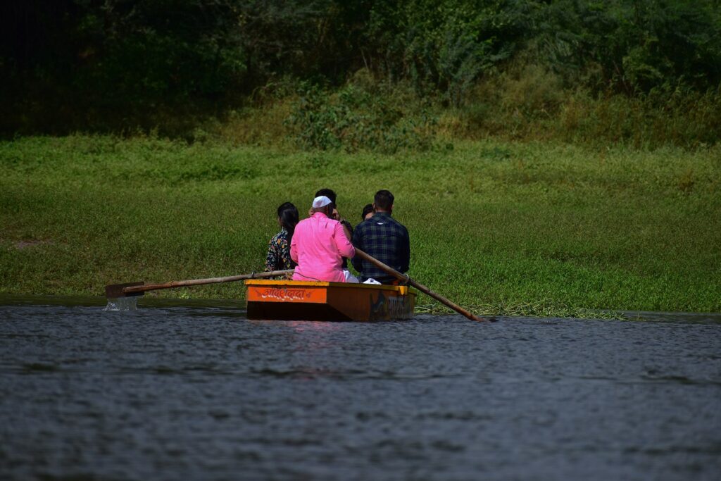 Four people in a boat, one in a pink shirt, paddle quietly on a calm river surrounded by lush green vegetation.