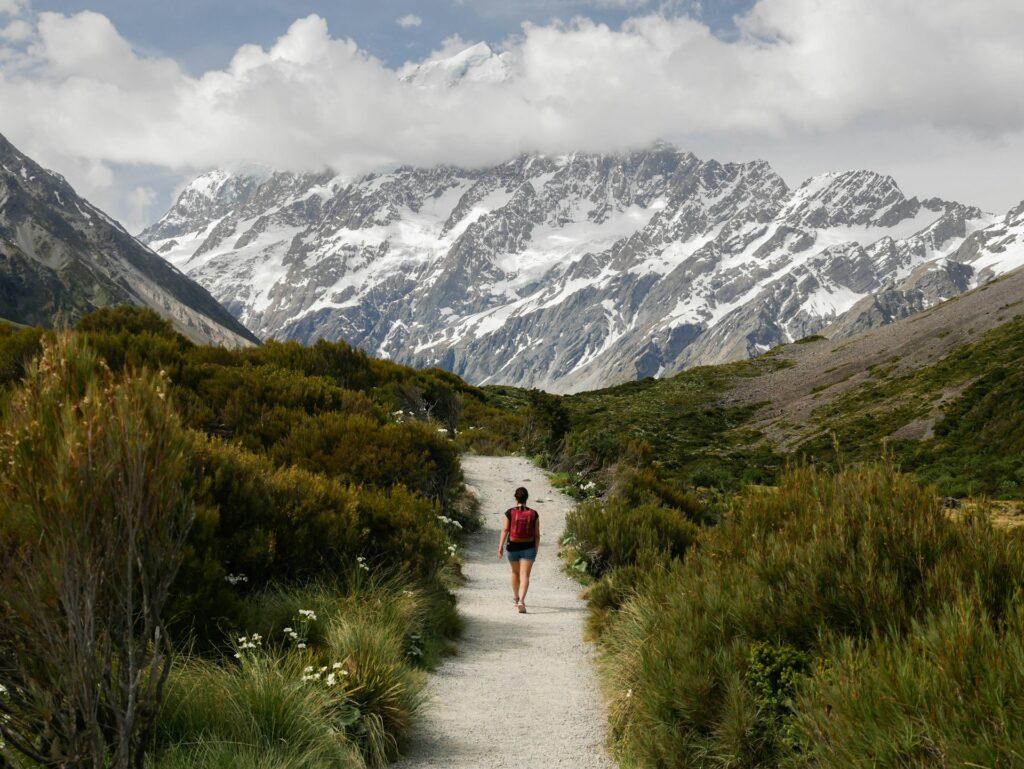 A hiker with a red backpack walks along a gravel trail, surrounded by lush greenery and towering snowy mountains under a cloudy sky.