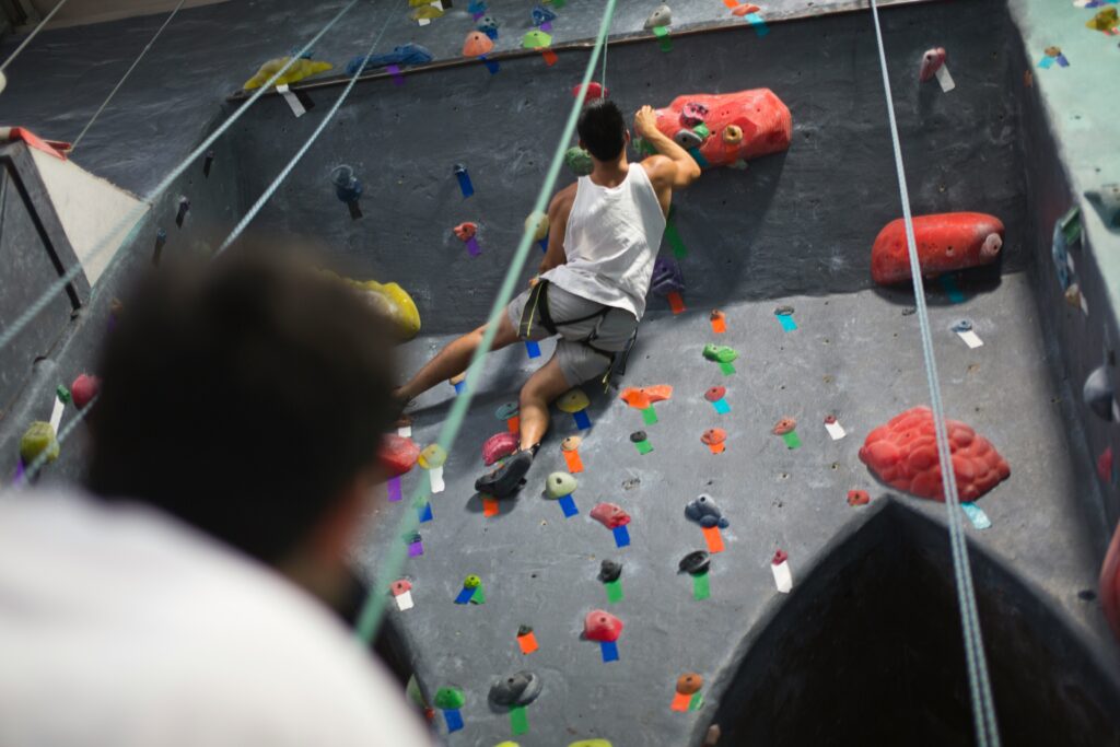 A climber in a tank top ascends a colorful bouldering wall, with a blurred figure observing from below. Bright holds are scattered on the wall.