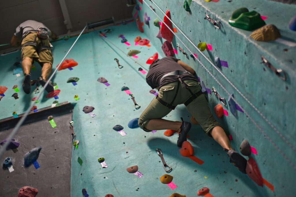 Two climbers ascend a vertical rock wall indoors, surrounded by colorful climbing holds and safety harnesses.