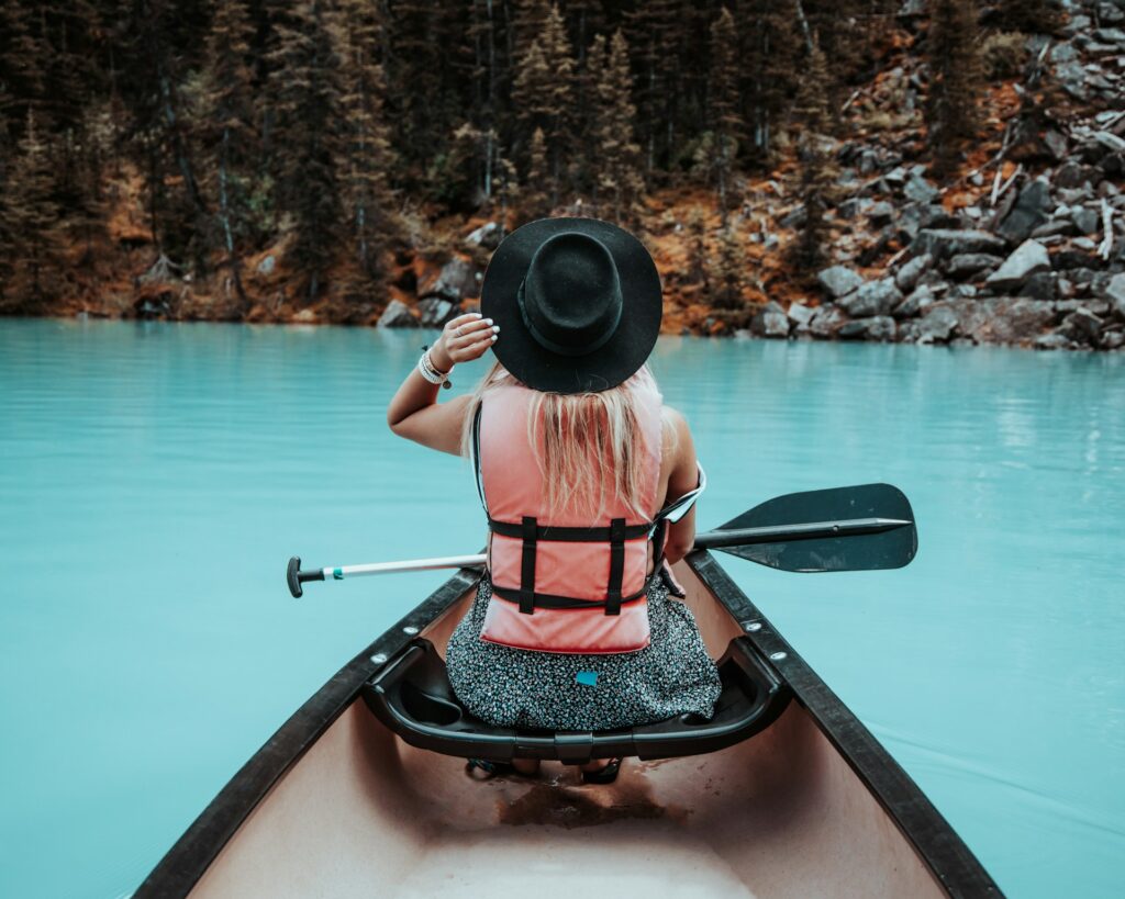 A woman in a pink life jacket and black hat sits in a kayak, paddling on tranquil turquoise waters surrounded by forested rocks.