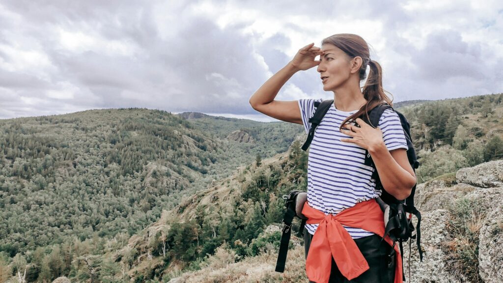 A hiker in a striped shirt and red jacket stands on a rocky outcrop, looking out over lush green hills under a cloudy sky.