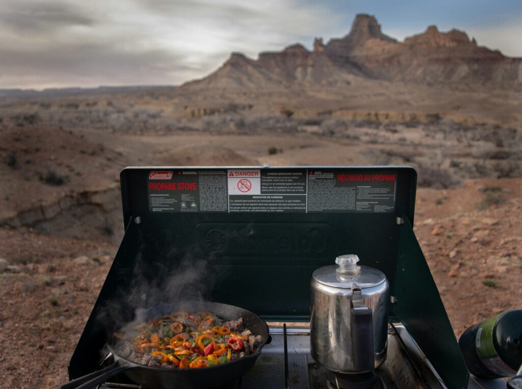 A propane stove with sizzling vegetables and meat steams in the foreground, set against a rugged desert landscape under a grey sky.