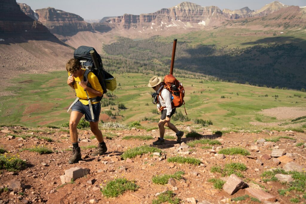 Two hikers ascend a rocky trail in a mountainous landscape, surrounded by green valleys and distant peaks under a clear sky.