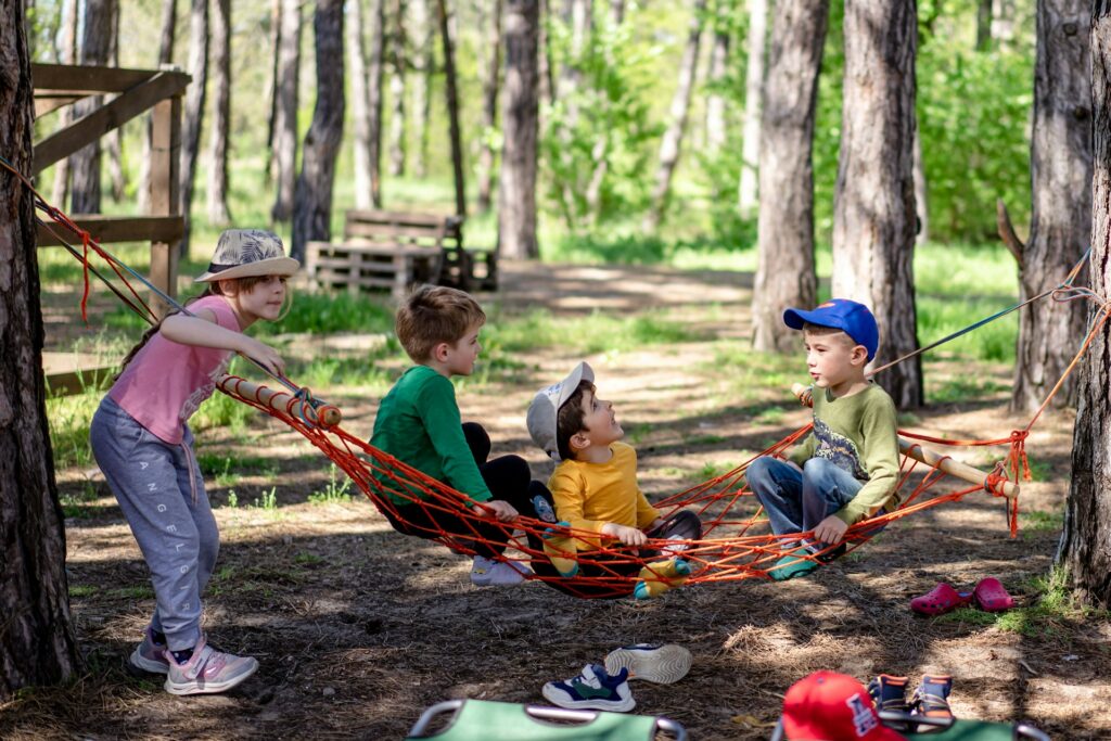Four children play on a colorful hammock in a sunlit forest, surrounded by trees, with a playful and cheerful atmosphere.