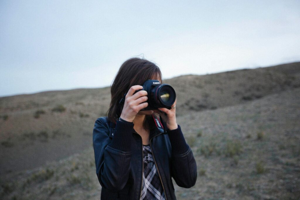 A woman wearing a black jacket stands outdoors, holding a Canon camera up to her eye, ready to capture the landscape.
