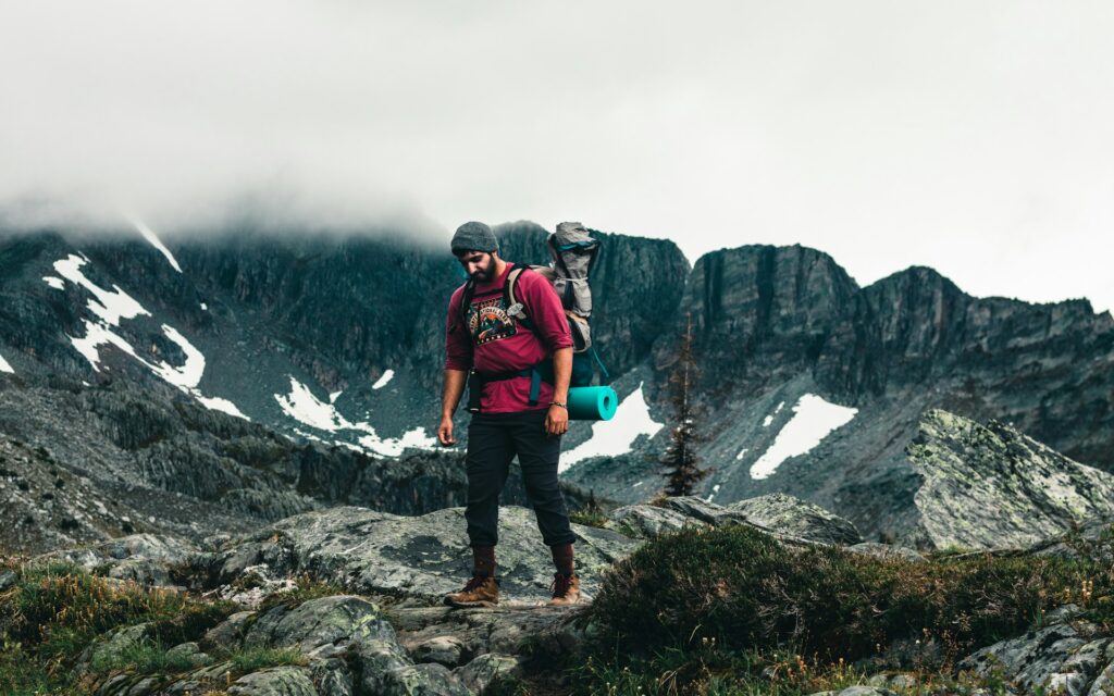 A hiker with a backpack stands on rocky terrain, surrounded by towering mountains and patches of snow under a cloudy sky.