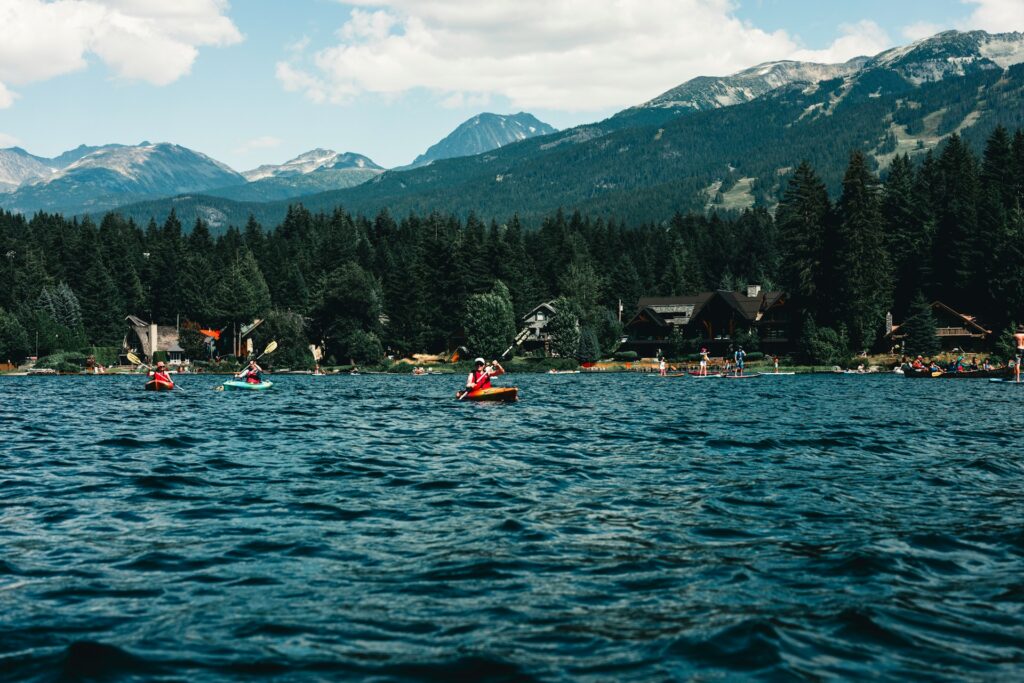 Two kayakers paddle on a serene lake surrounded by lush forests and mountains, with people enjoying various water activities nearby.