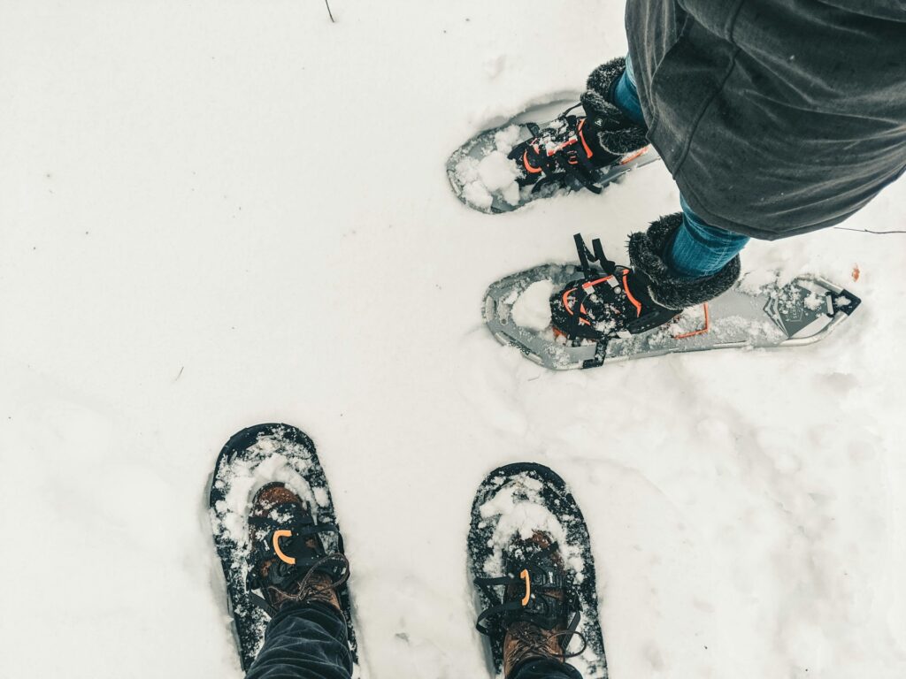 Two pairs of snowshoes in freshly fallen snow, one brown boot and one blue boot visible, surrounded by a white snowy landscape.