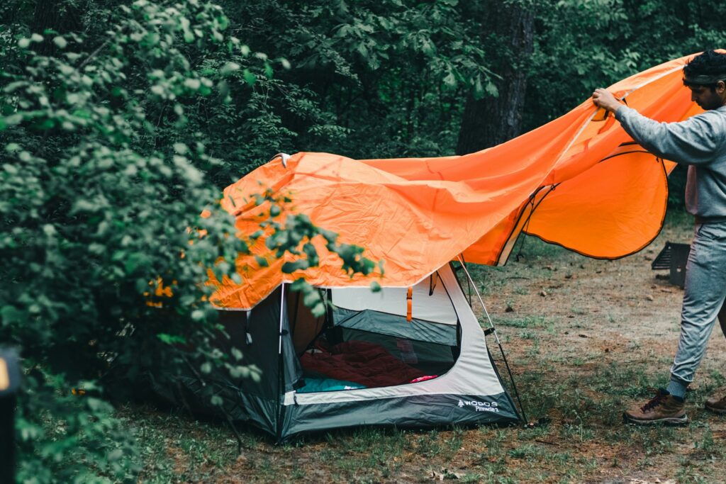 A person secures an orange tarp over a camping tent nestled in a lush forest, enhancing protection from the elements.