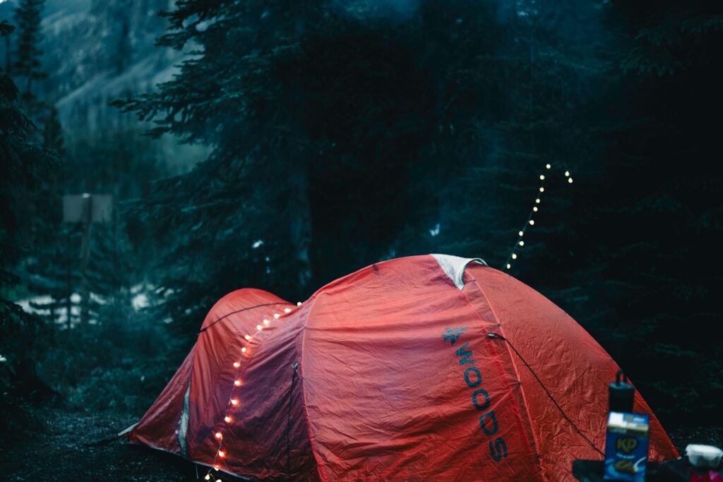 A red camping tent nestled among dark trees, adorned with string lights, evoking a cozy, outdoor camping atmosphere.