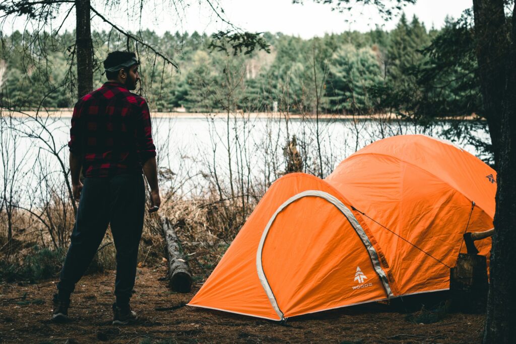 A person in a red and black flannel stands near an orange tent by a serene lake, surrounded by tall trees and nature.