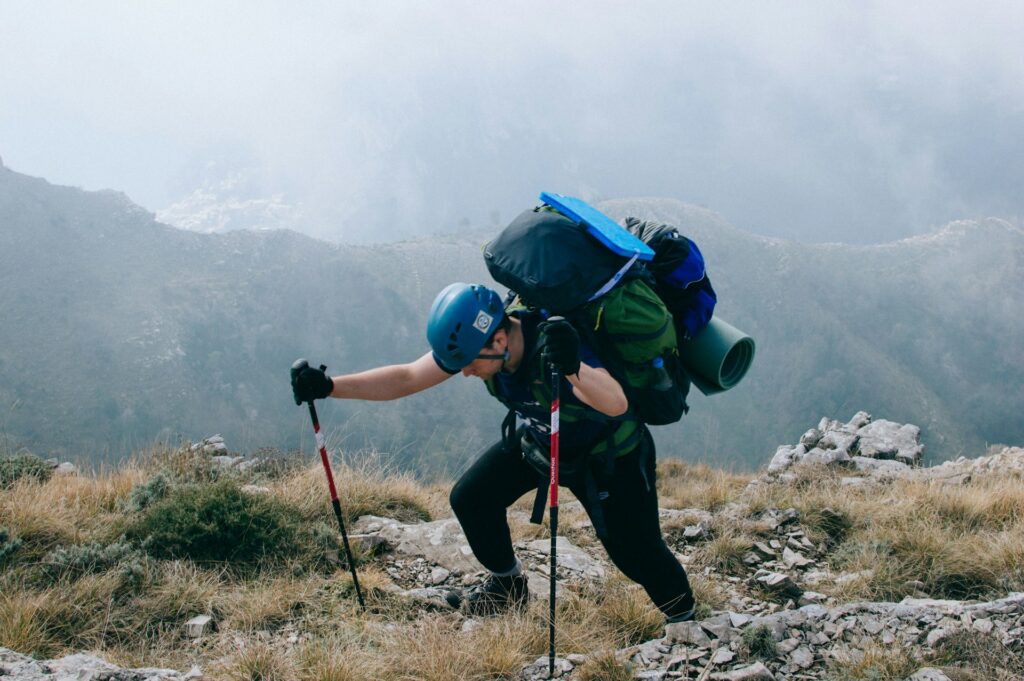 A climber ascends a rocky, grassy trail with hiking poles, a heavy backpack, and a blue helmet, surrounded by misty mountains.