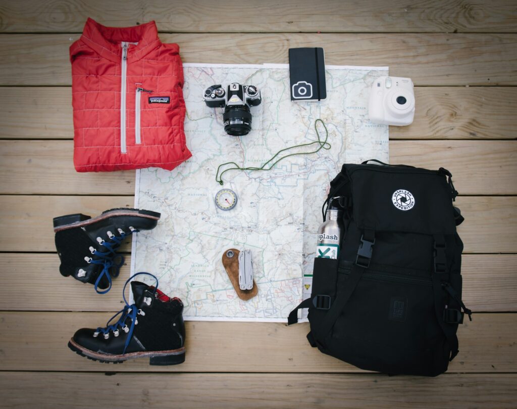 A flat lay of outdoor gear: a red jacket, cameras, a map, compass, black boots, a backpack, and a water bottle on wooden flooring.
