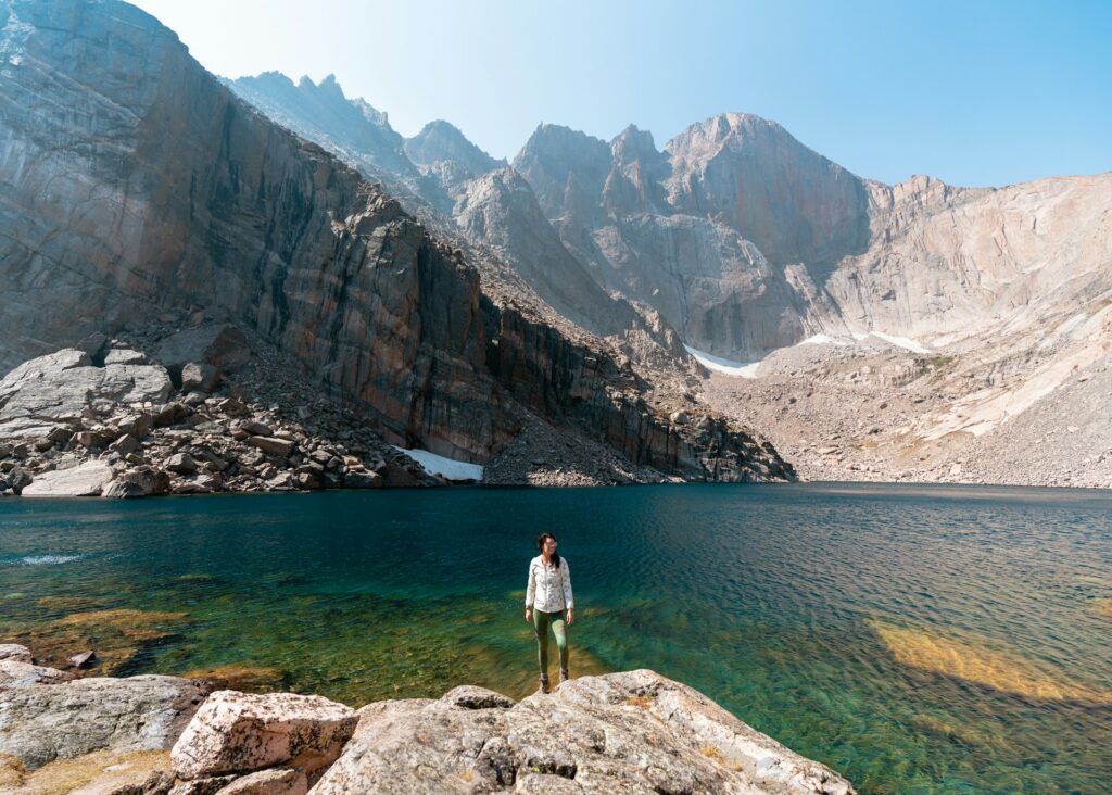 A hiker stands at the edge of a turquoise lake surrounded by towering rocky mountains under a clear blue sky.