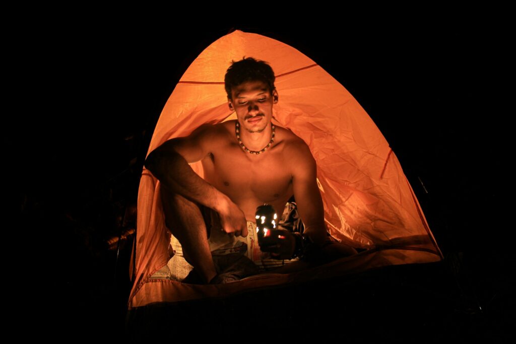 A person sitting cross-legged inside an orange tent, holding a flashlight, in a dark outdoor setting.