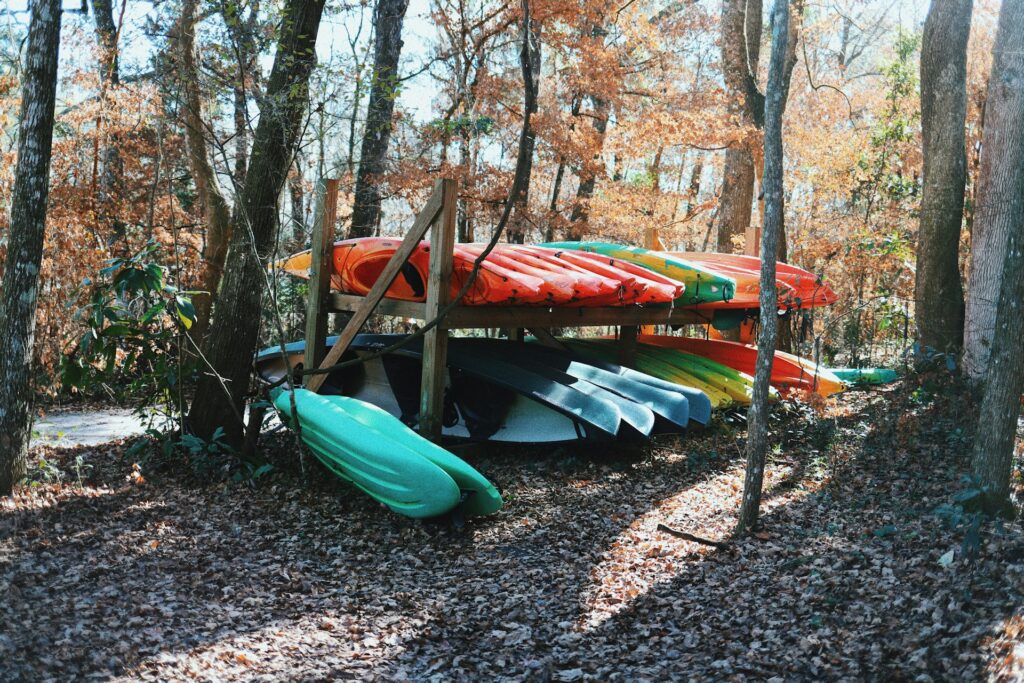 A colorful stack of kayaks resting on wooden racks among autumn trees and fallen leaves in a serene forest setting.