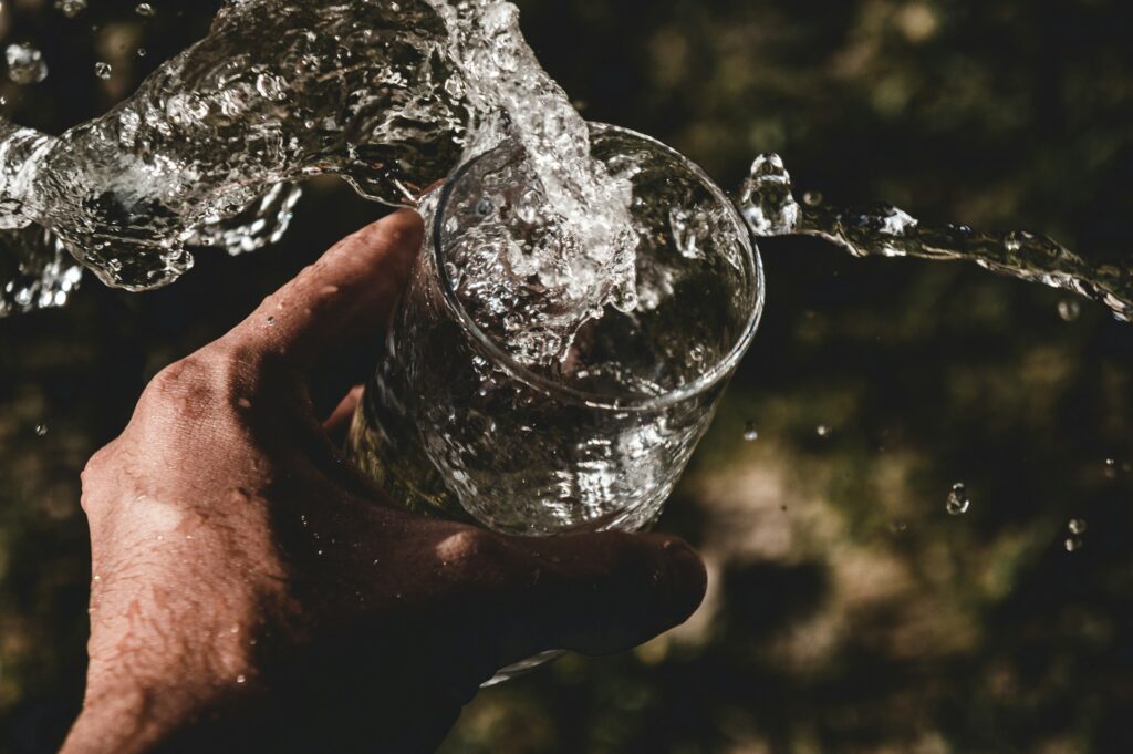 A hand holds a clear glass as water pours rapidly into it, creating splashes and droplets against a soft, blurred background.