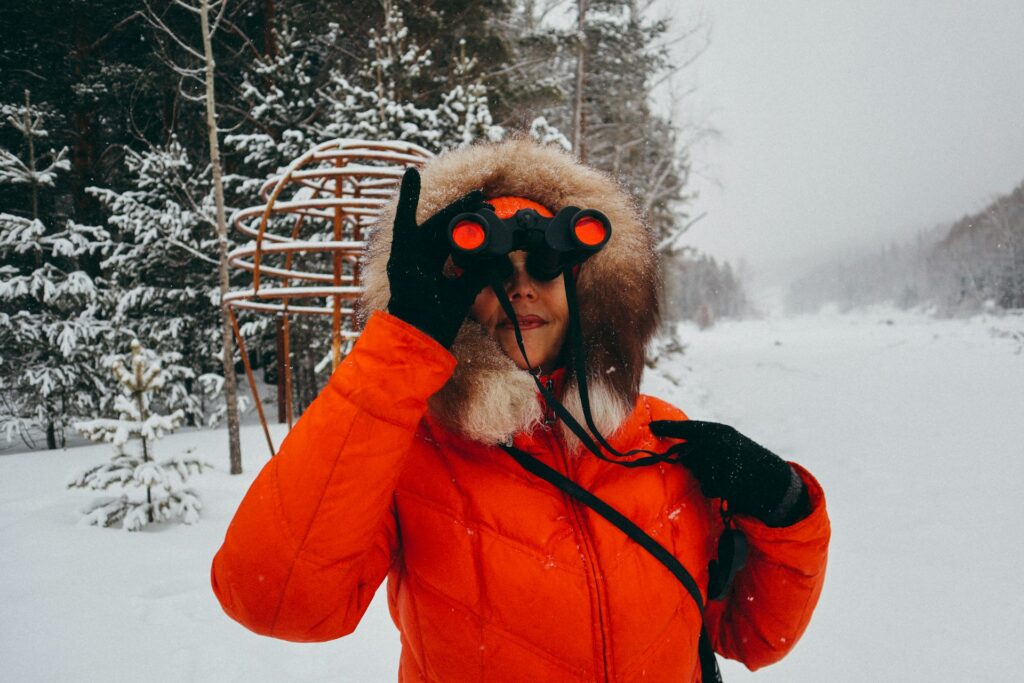 A person in an orange parka with a fur-lined hood stands in a snowy landscape, holding binoculars, surrounded by tall trees.