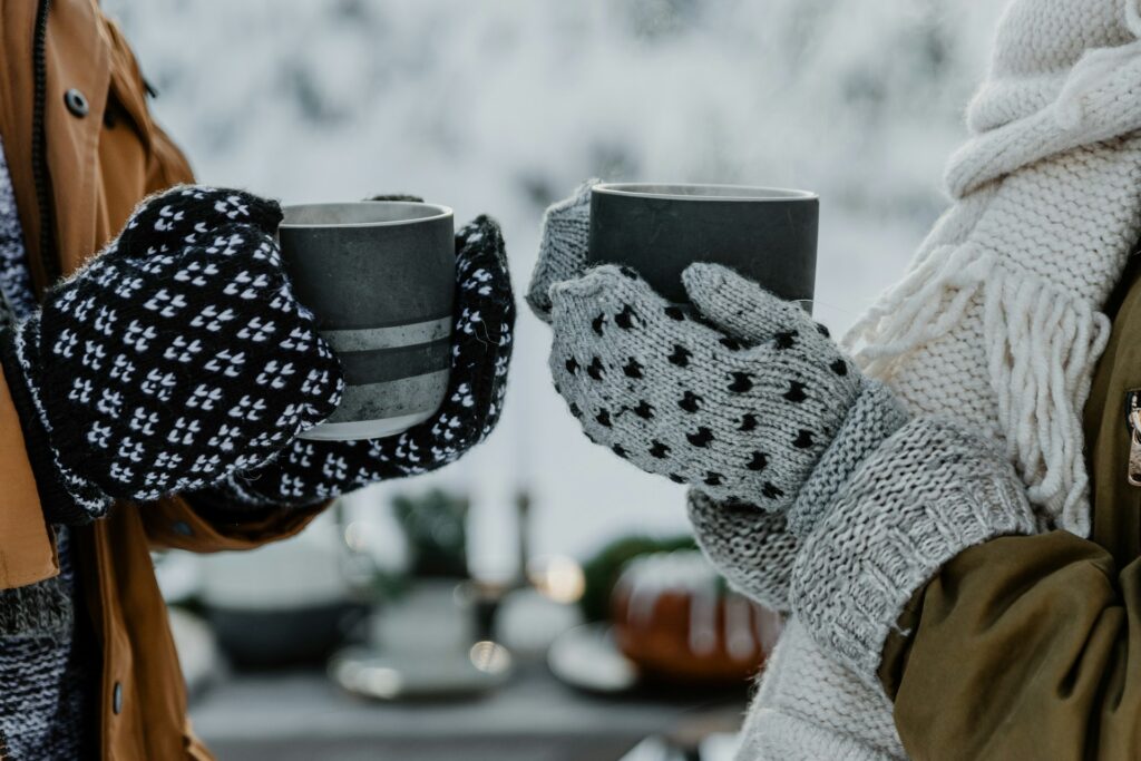 Two people wearing cozy mittens hold steaming cups in a snowy outdoor setting, surrounded by festive winter decor.