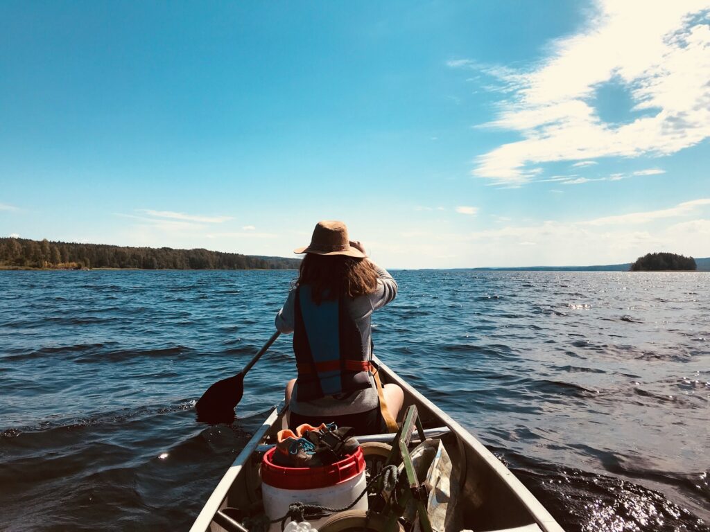 A person paddles a canoe on a serene lake, wearing a wide-brimmed hat under a bright blue sky with scattered clouds.