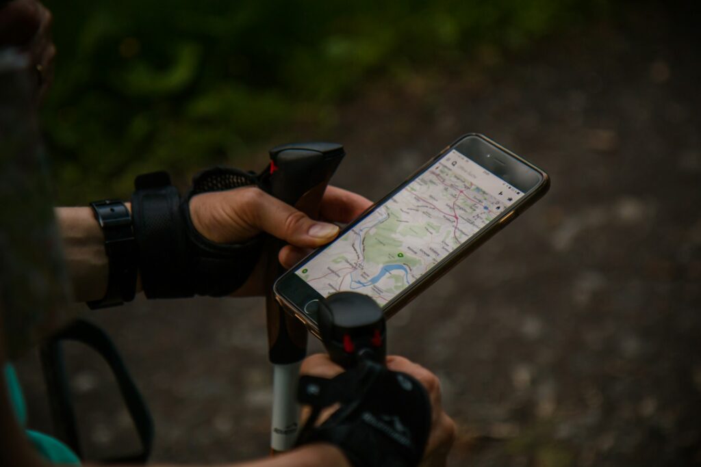 A hiker checks a smartphone displaying a map while holding a trekking pole, surrounded by lush greenery on a trail.