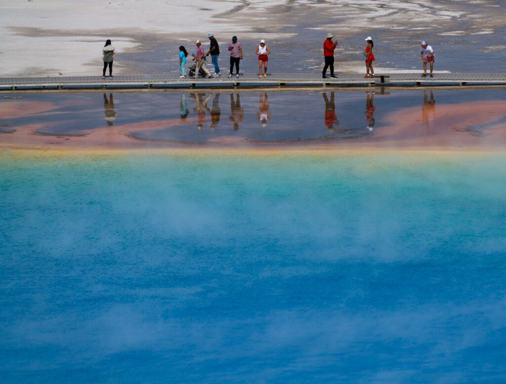 Visitors stand on a boardwalk near a vibrant, multicolored hot spring, reflecting the blue and orange hues in the water.