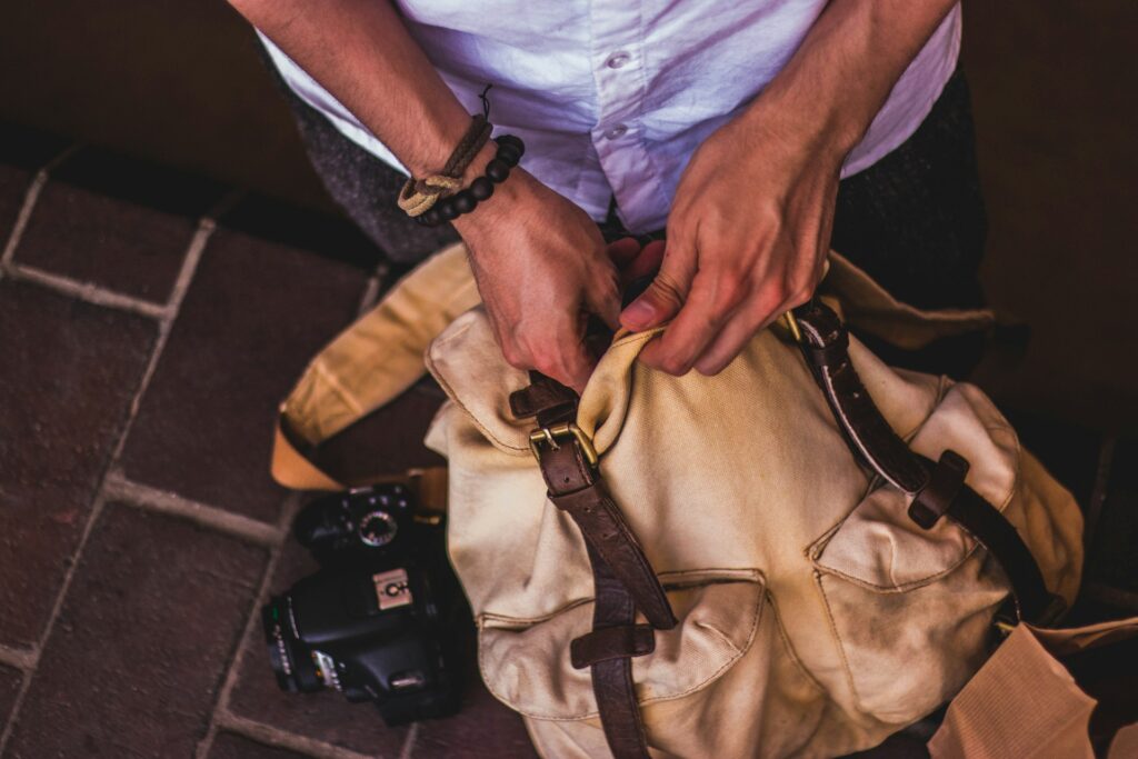 A person’s hands are seen opening a beige canvas bag with brown leather straps, next to a black camera on the ground.