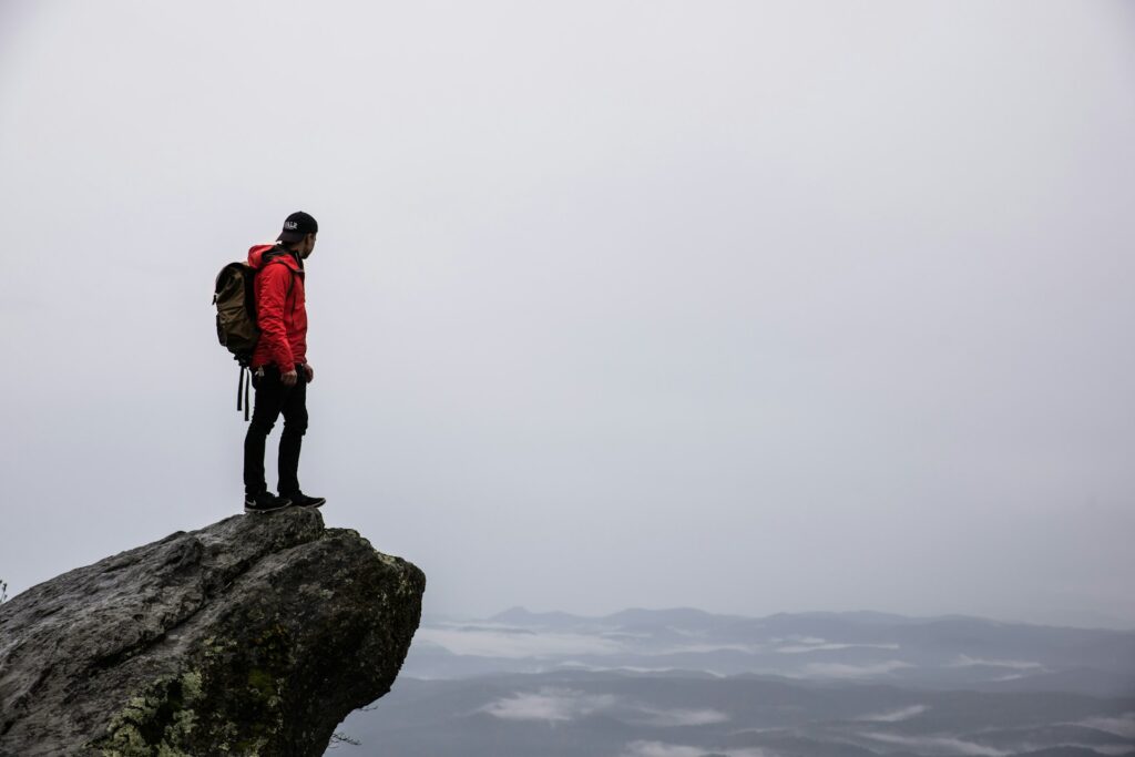 A hiker in a red jacket stands atop a rocky outcrop, gazing at a misty mountain landscape under a cloudy sky.
