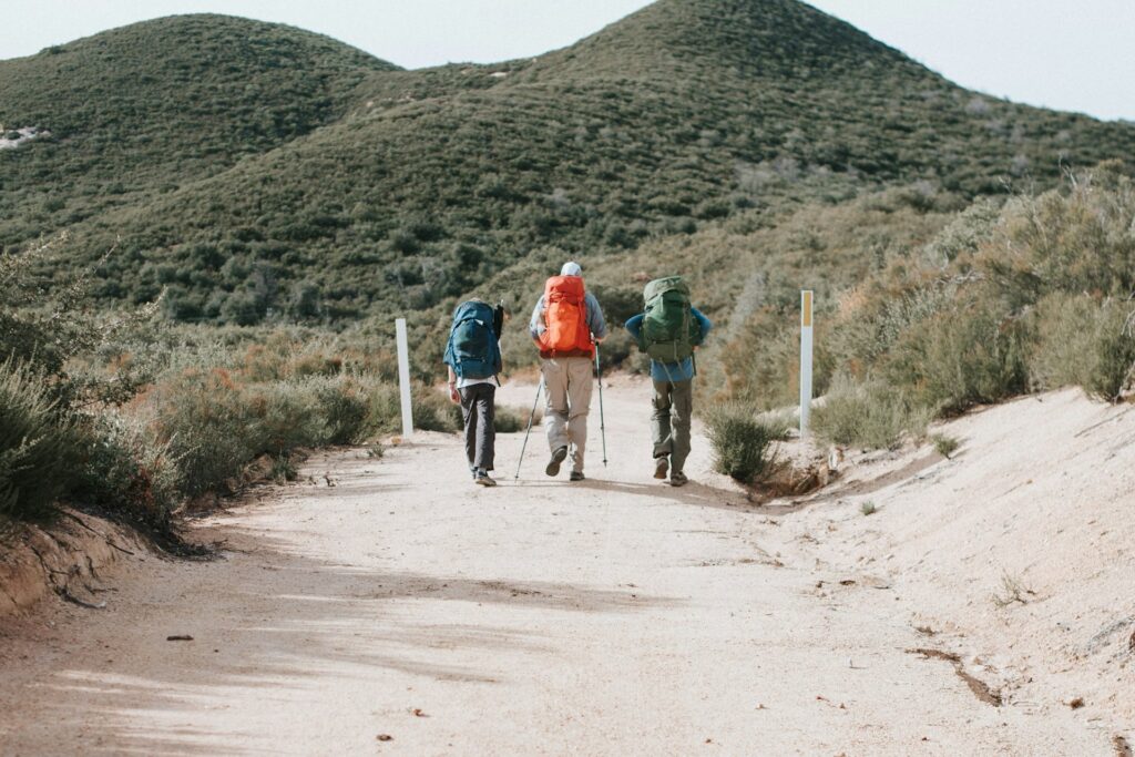 Three hikers with backpacks walk along a sandy path, flanked by green hills and shrubs, under a bright sky.