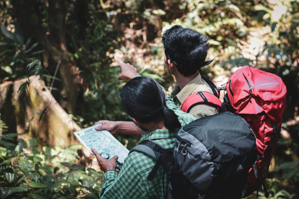 Two hikers in a lush forest, one pointing ahead while the other studies a map, both equipped with backpacks.