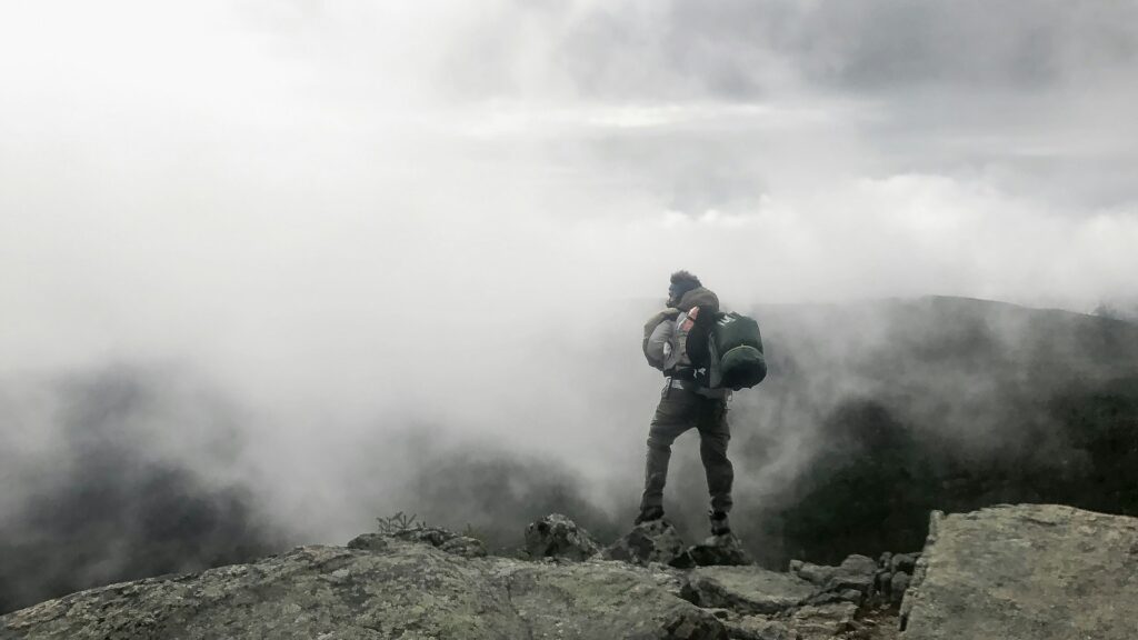A hiker stands on a rocky outcrop, gazing into a misty valley shrouded in clouds, embodying adventure and solitude.