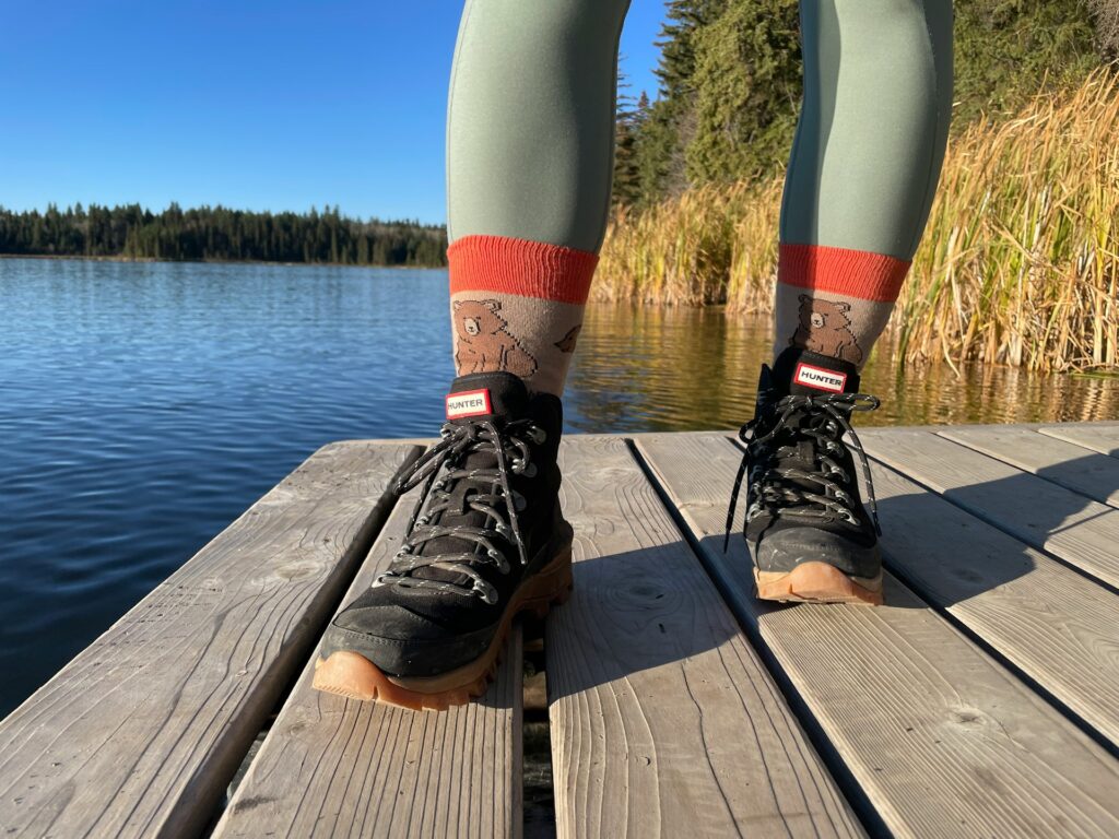 A close-up of hiking boots and playful bear-patterned socks on a wooden dock by a serene lake, surrounded by tall grass and trees.