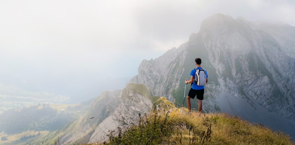 A hiker stands on a mountain summit, overlooking misty valleys and rugged peaks, with a backpack and trekking pole in hand.