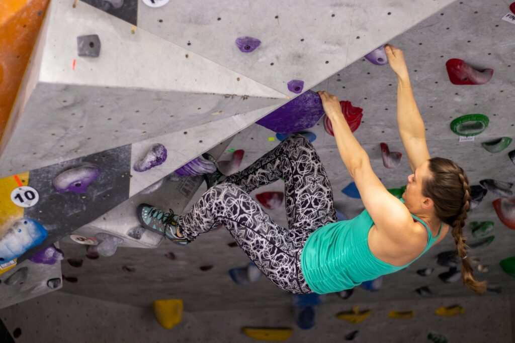 A climber in a green tank top and patterned leggings maneuvers on a bouldering wall with various colored holds.