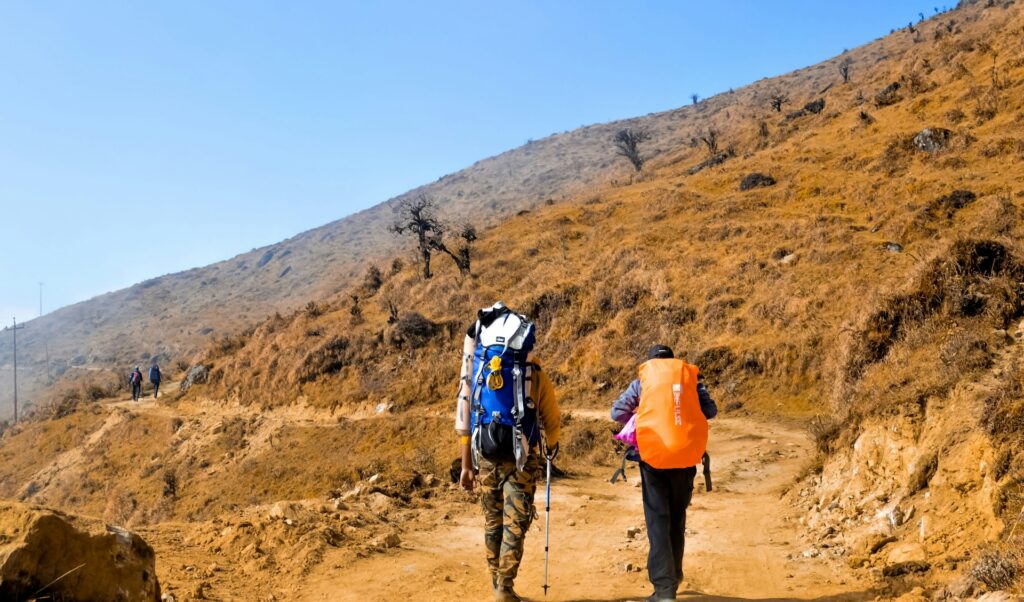 Two hikers trek a dusty path uphill, one carrying a large backpack and the other wearing an orange vest, amid dry, grassy terrain.